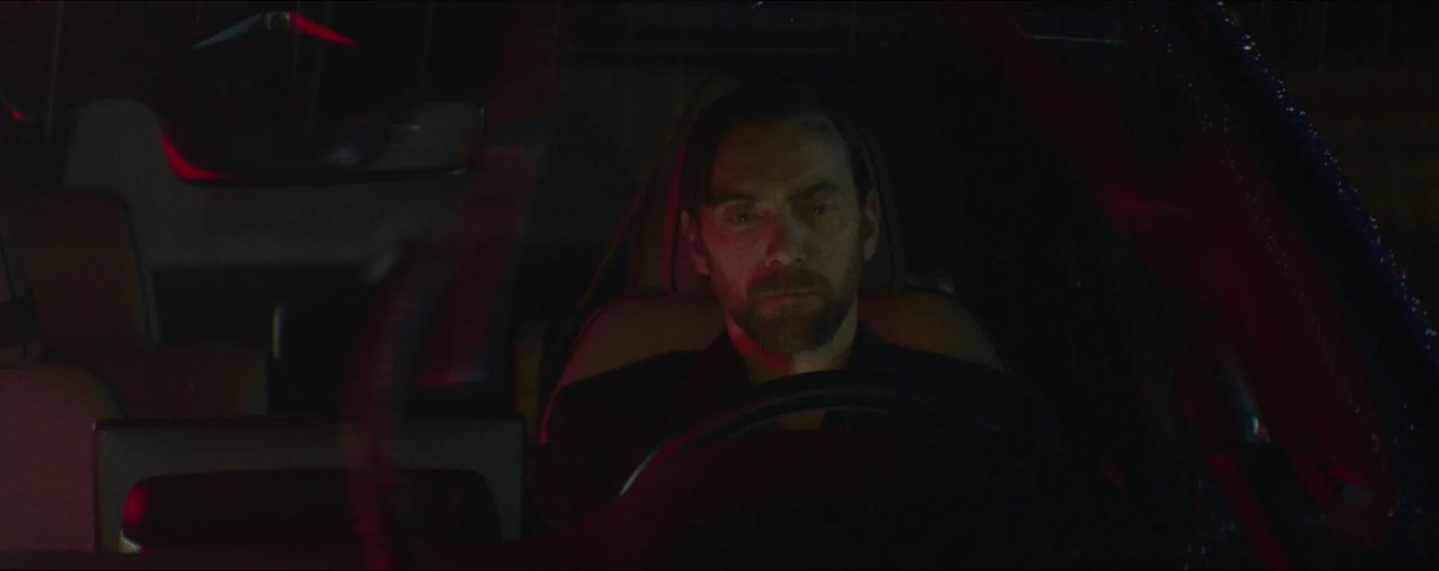 A man sits alone behind the wheel of a parked car, lit red from outside