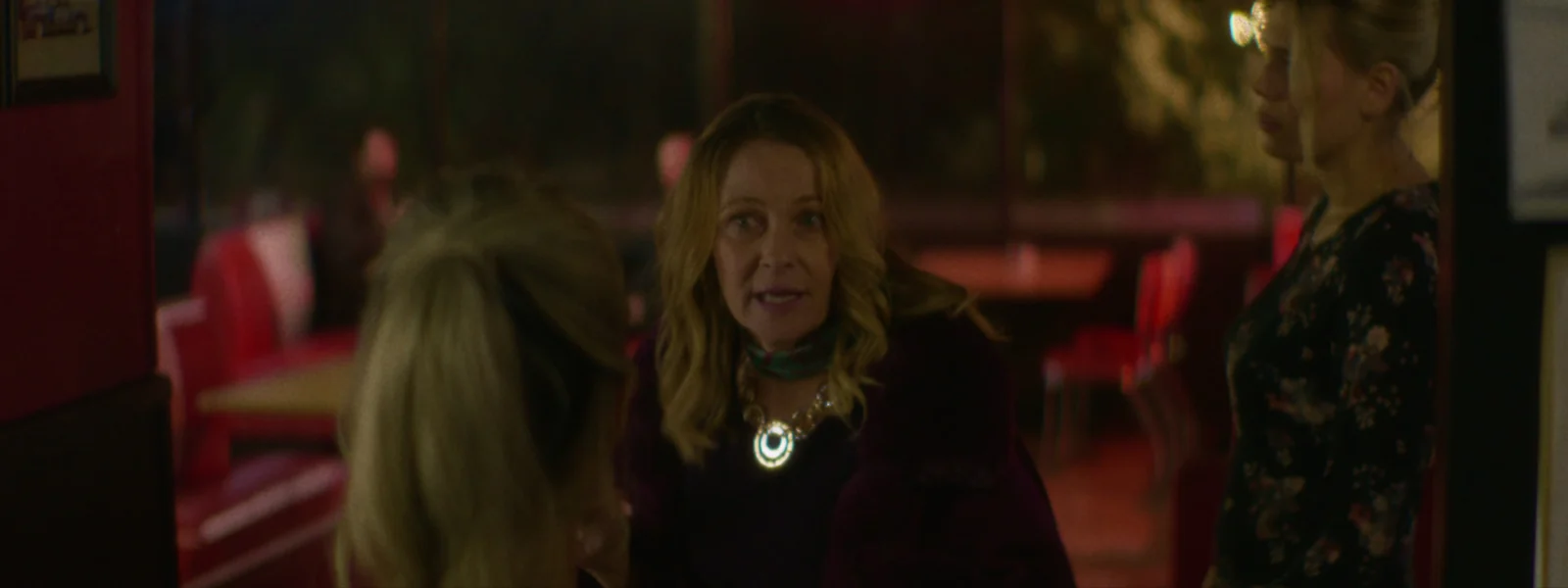 A woman in a purple coat stands over a booth mid-sentence