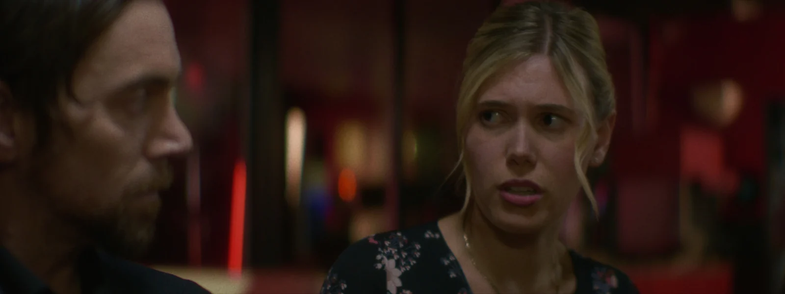 A young woman looks off past a man’s shoulder in a dim diner booth