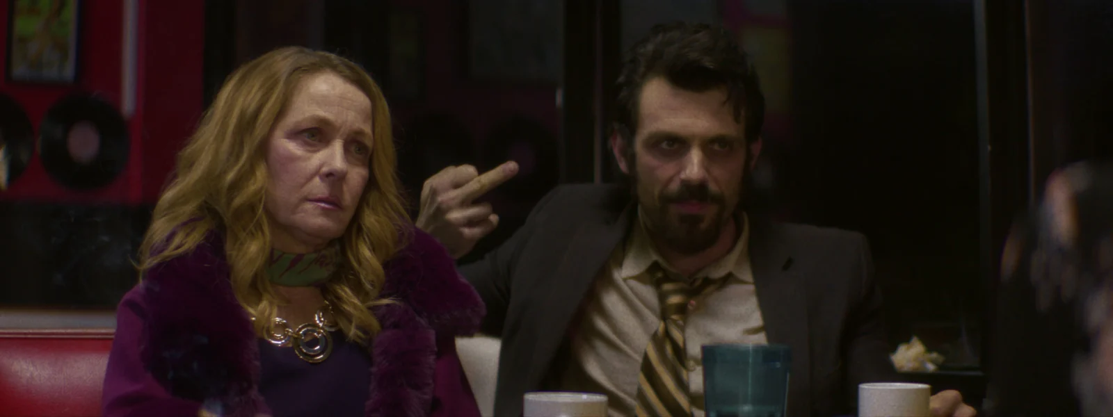 A woman in a purple fur coat and a man in a suit sit together in a diner booth
