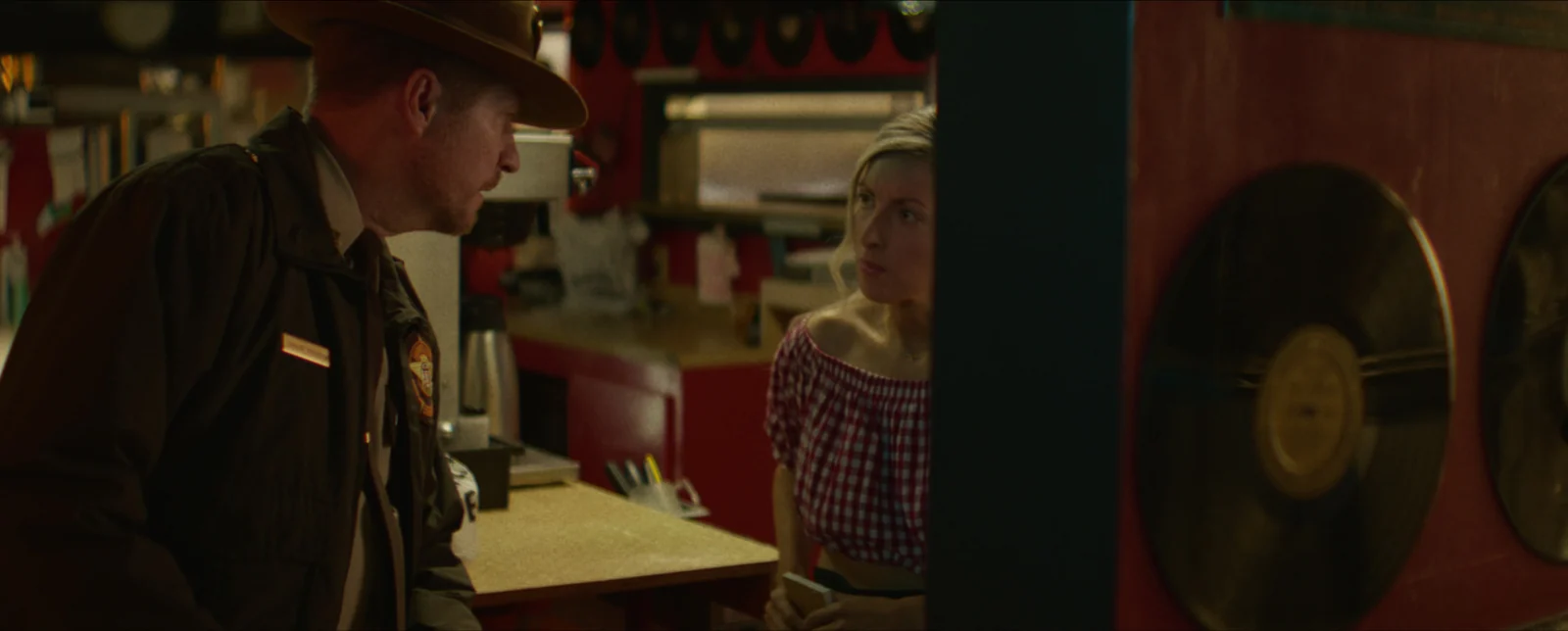 A park ranger leans on the counter talking to a waitress, gold records on the wall behind