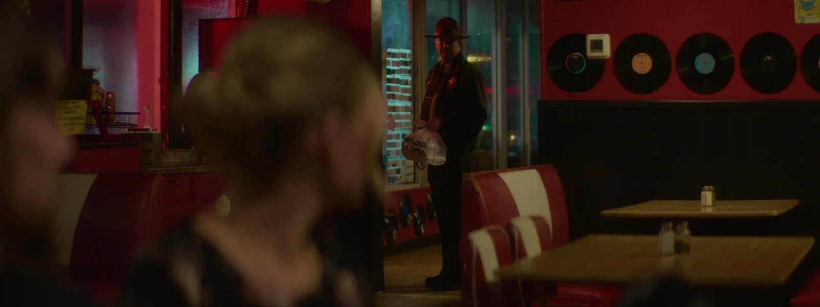 A ranger stands at the door of a near-empty diner, seen past a woman in the foreground