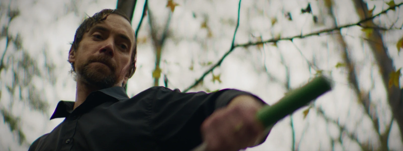 A man raises a long-handled tool, seen from below against bare winter branches