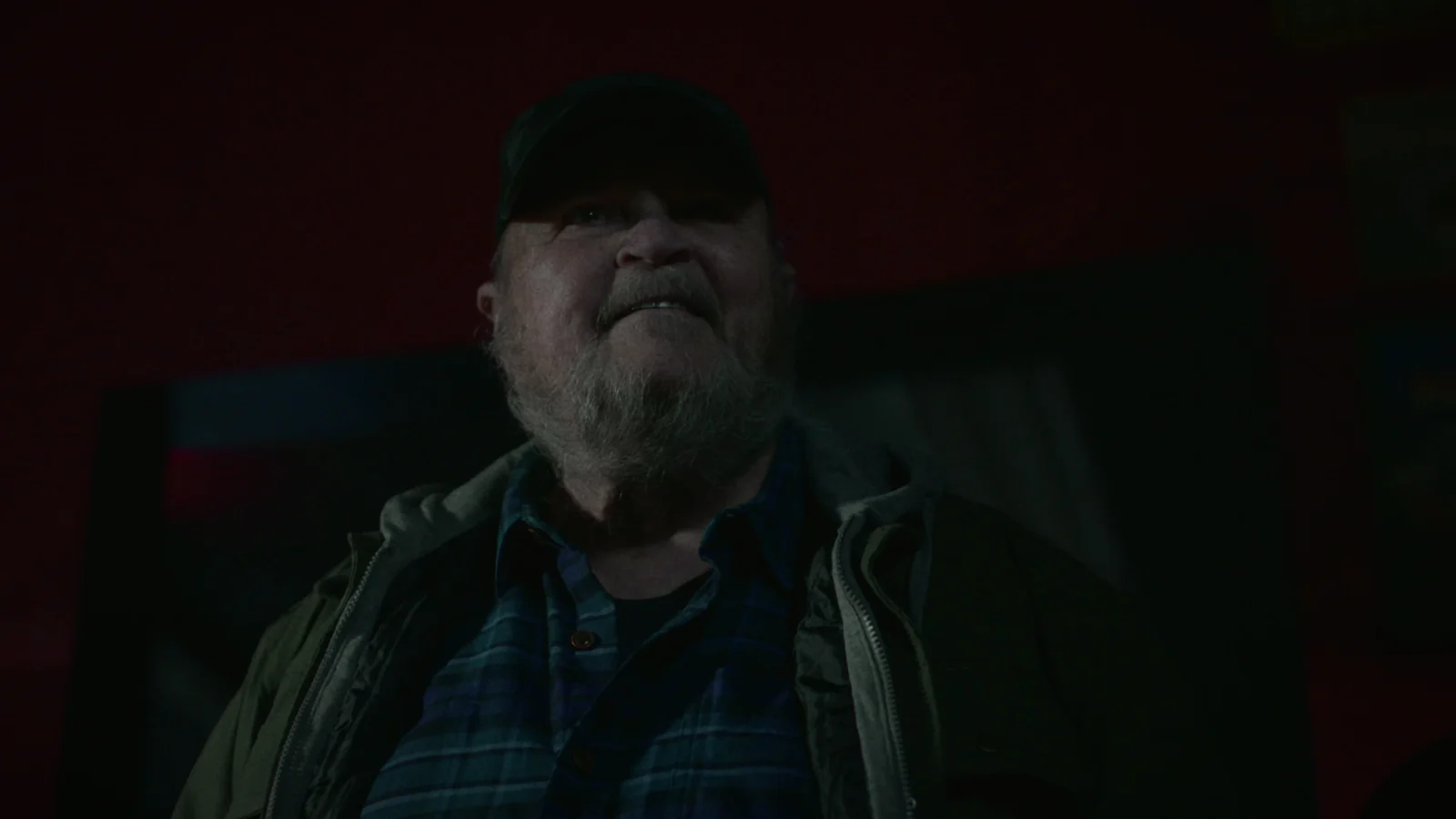An older bearded man in a cap, lit deep red, staring ahead